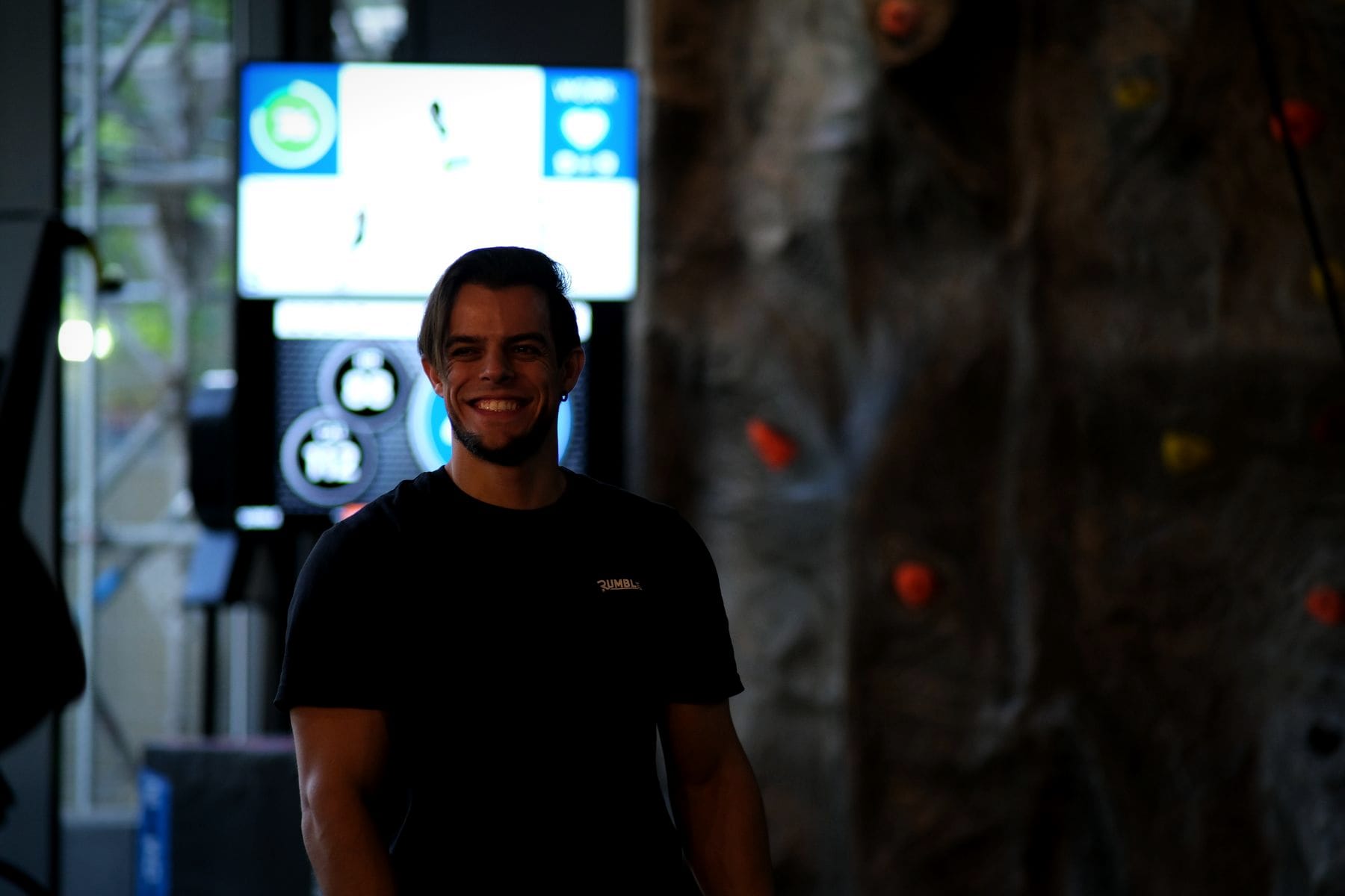 A smiling coach on the circuit floor, with the workout timer on the screen behind him