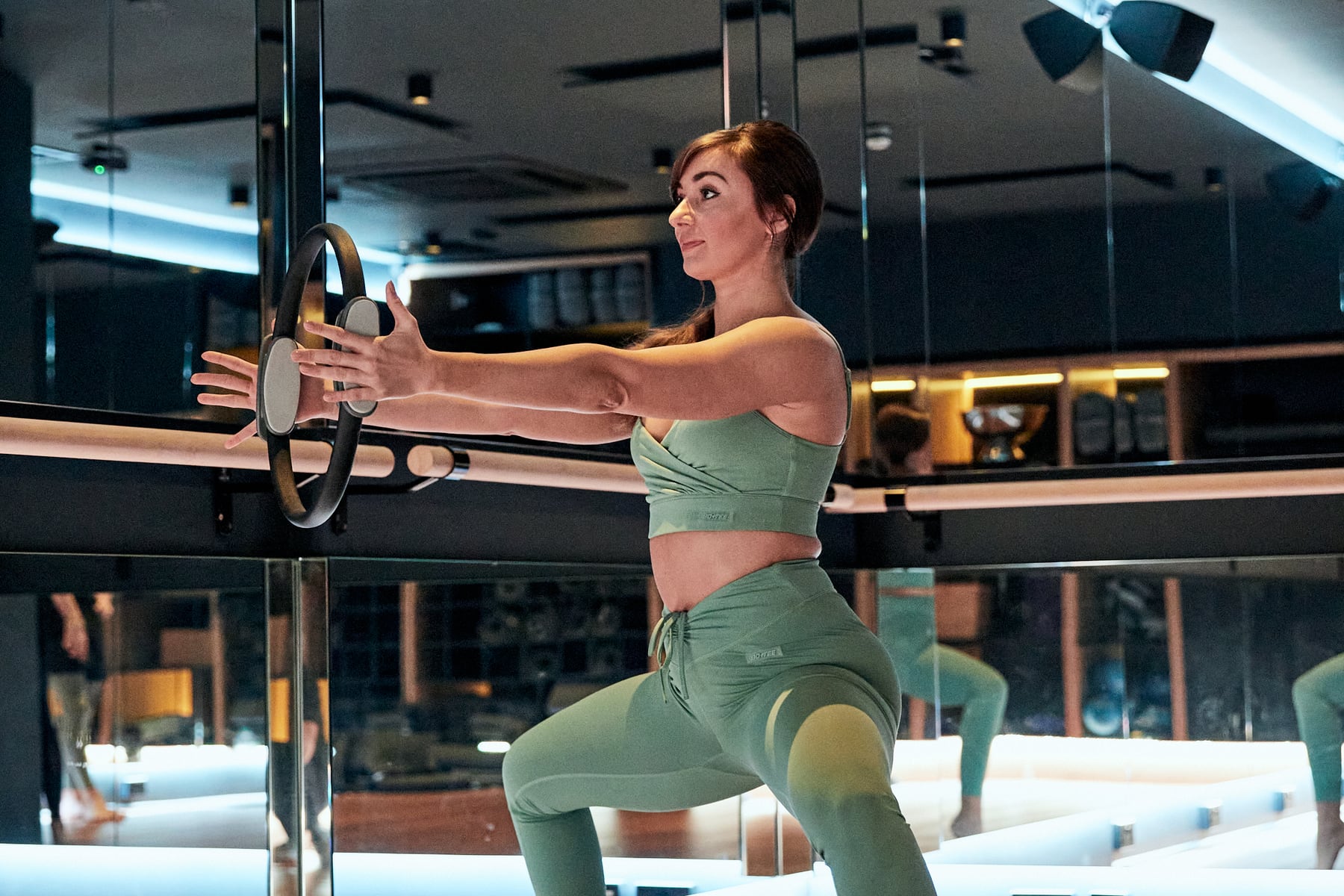 A woman holds a wide plié at the barre, pressing a Pilates ring between her hands, in the Mind & Body studio
