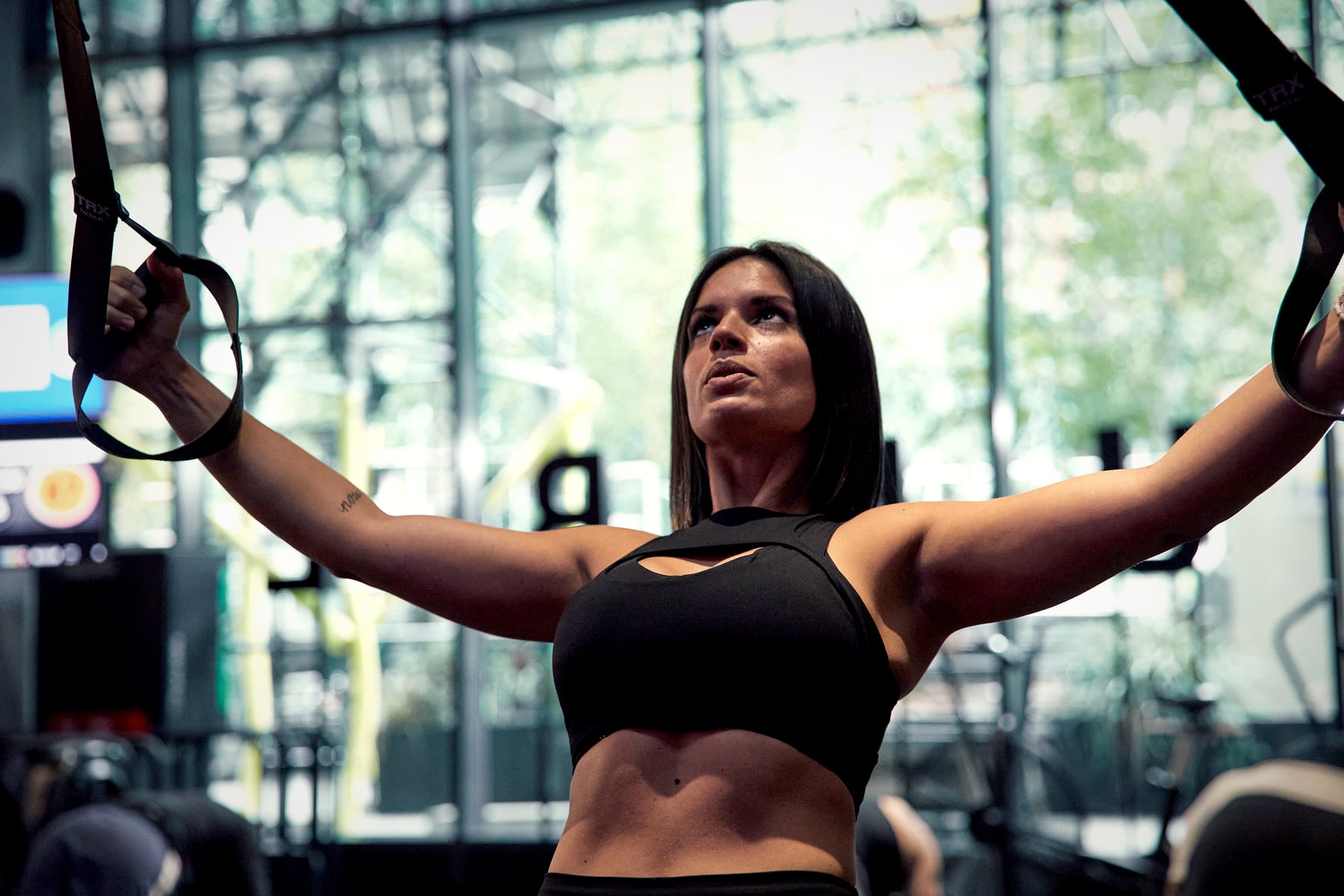 A woman pulls on suspension straps, arms spread wide, during a circuit class at Rumble’s Dalston studio, with the big front windows behind her